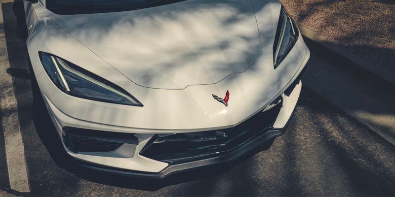 White Chevrolet Corvette Stingray parked on the street next to a tree as the sun casts a shadow of the tree on the hood